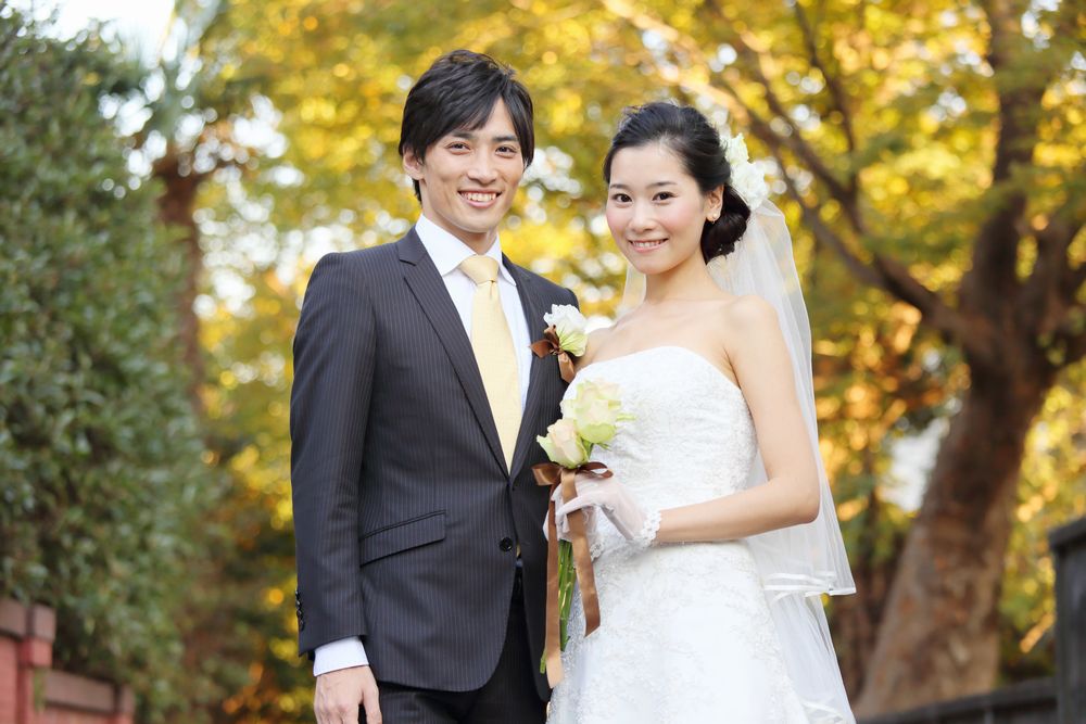 紅葉した木々を背景に、笑顔で並んで立つウェディング姿の男女